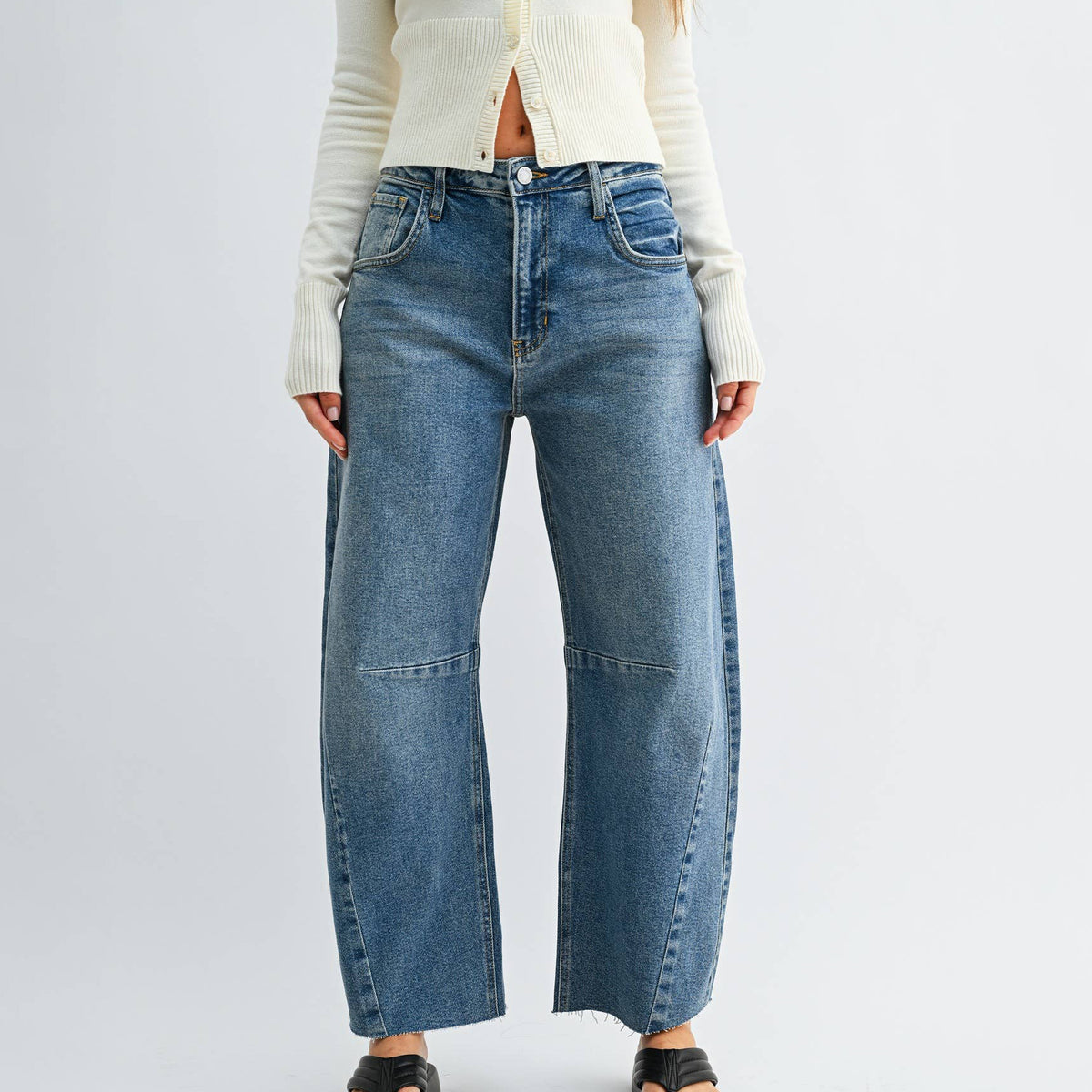 A model wearing a pair of wide-leg blue denim jeans with distinctive side seam details and a raw hem, styled with a cream ribbed cardigan and black sandals against a light gray studio background.