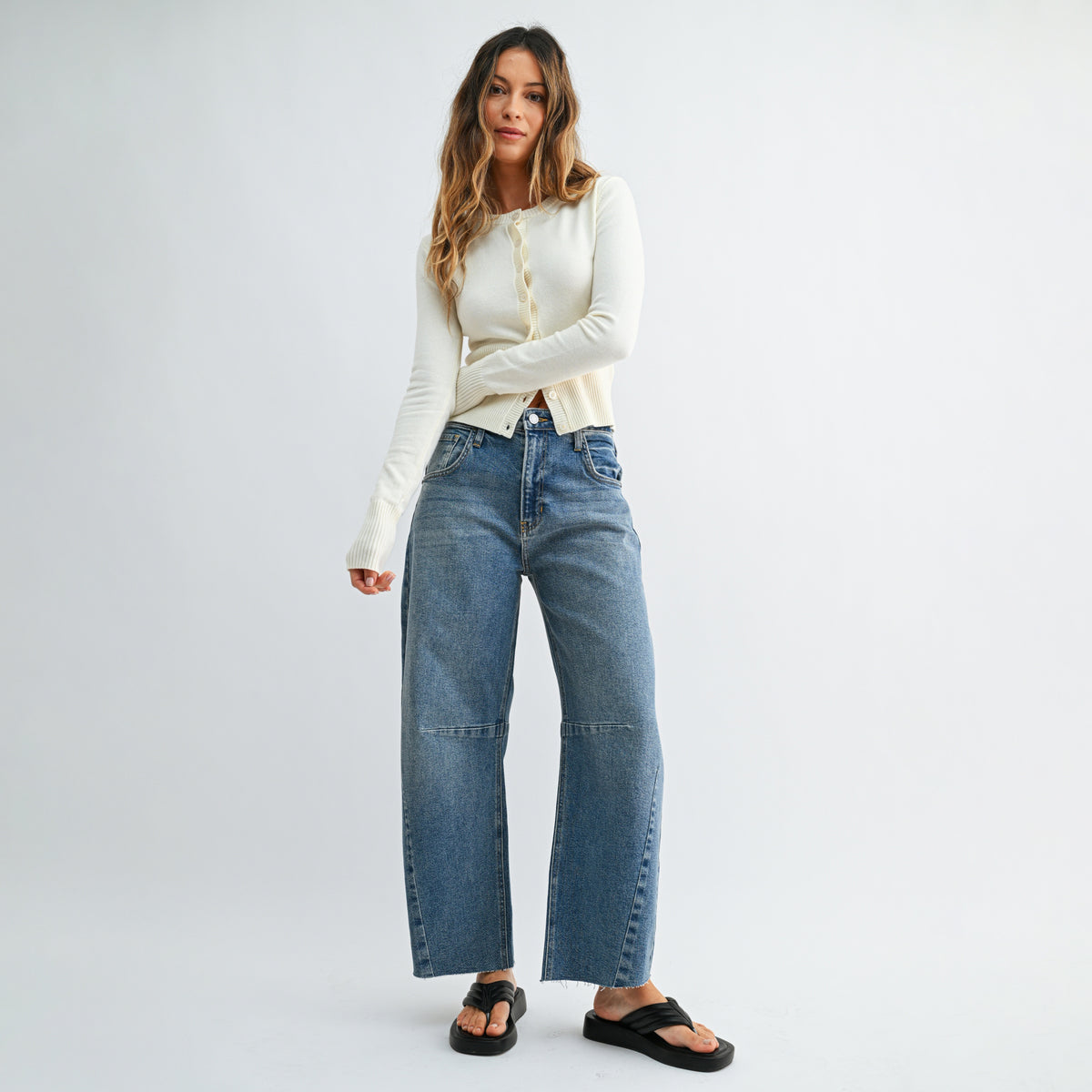 A woman modeling a pair of wide-leg blue denim jeans with a cream ribbed long-sleeve top and black slide sandals, standing against a light gray studio background.
