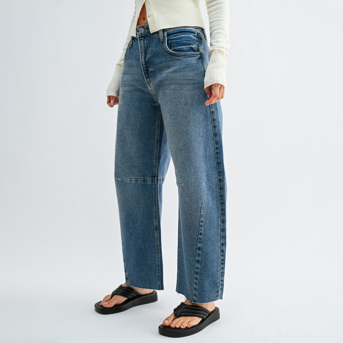 A model wearing a pair of wide-leg blue denim jeans with distinctive seam detailing down the sides, styled with a cream ribbed sweater and black slide sandals against a light gray studio background.