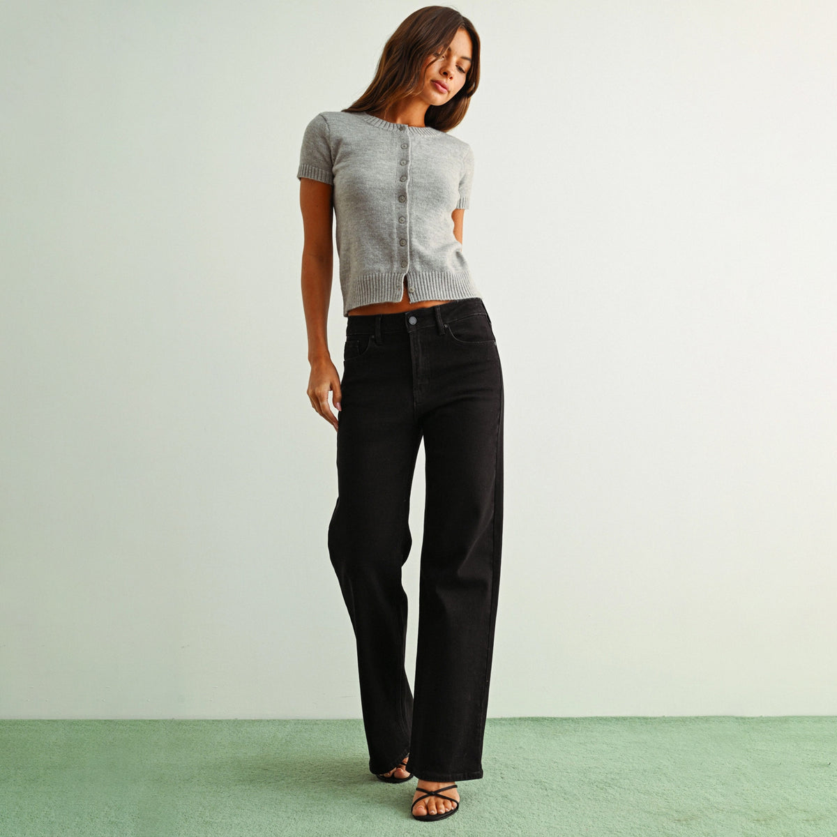 A woman modeling a light gray short-sleeve cropped knit top and black wide-leg jeans against a pale green wall and green floor.