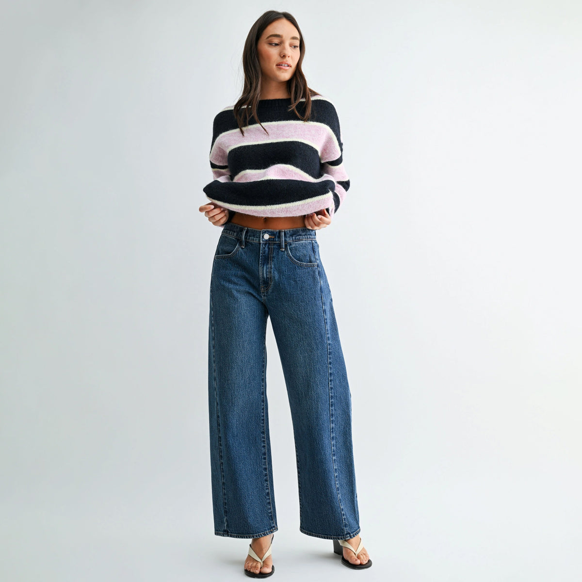 A woman modeling a black and light pink striped cropped knit sweater paired with high-waisted, wide-leg blue jeans and strappy heels, set against a plain light gray studio background.