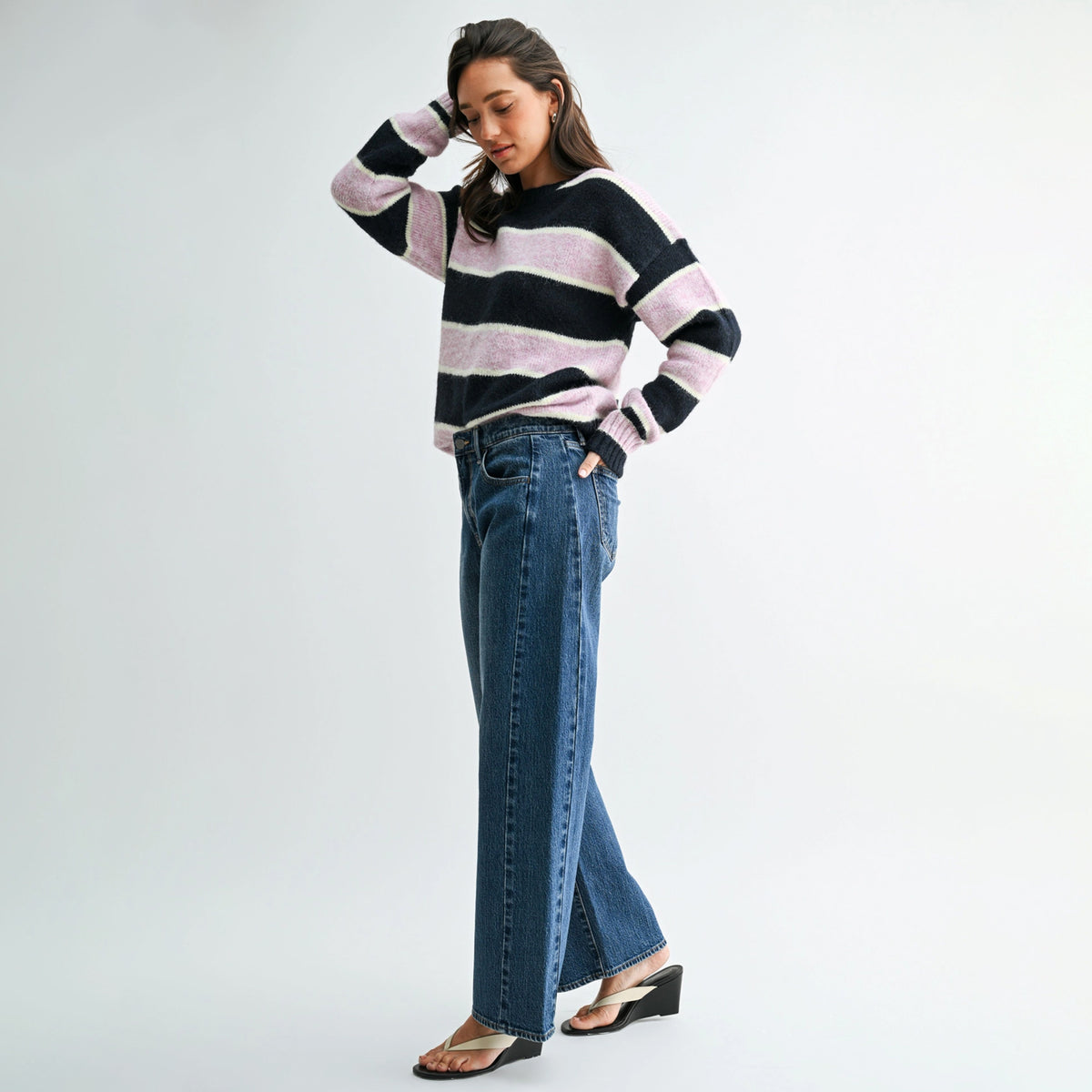 A woman modeling a fuzzy knit sweater with horizontal black and light pink stripes, paired with wide-leg blue jeans and black wedge sandals, against a plain light gray studio background.