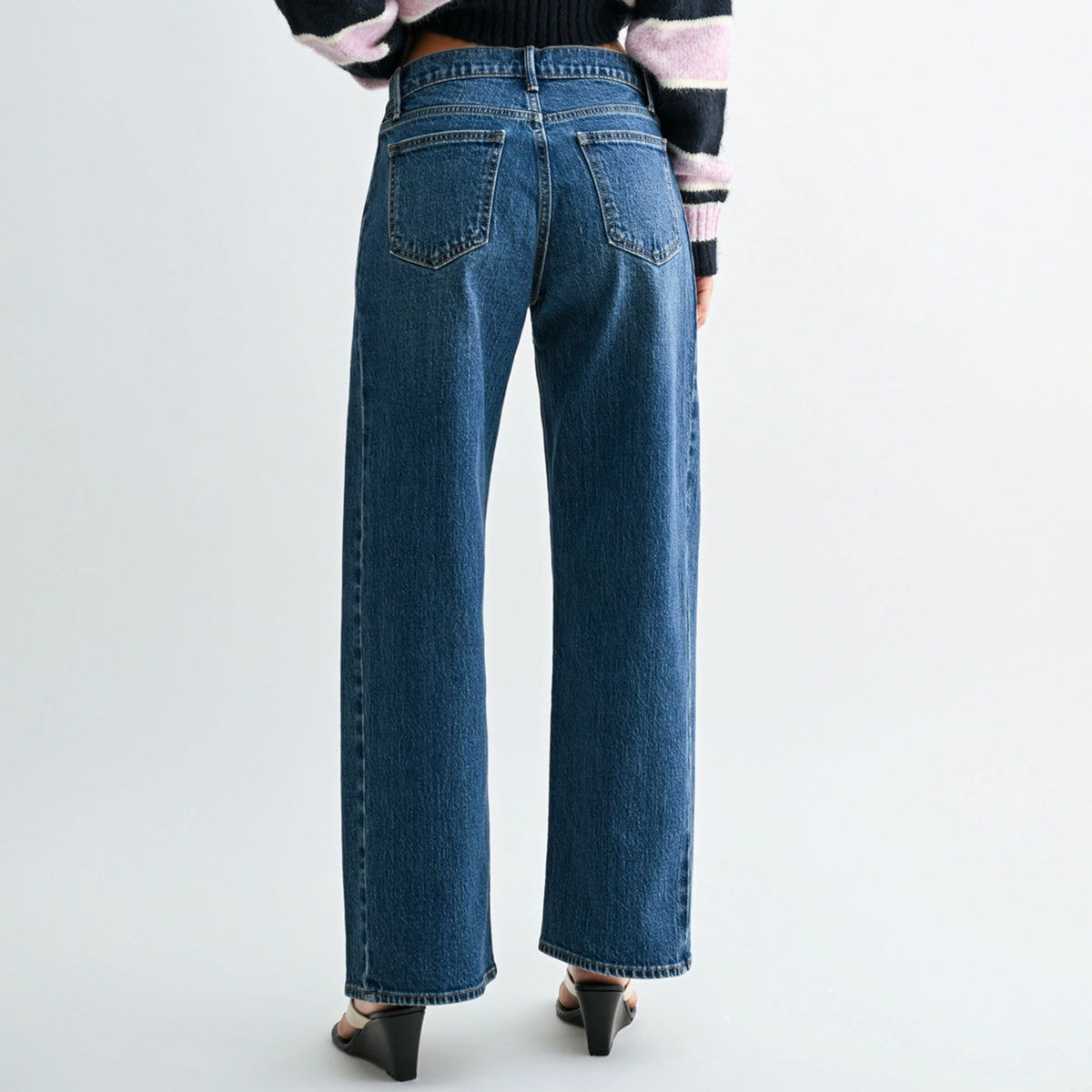 A person shown from the back wearing dark blue wide-leg denim jeans, a black and pink striped sweater, and black heels against a plain light gray studio background.