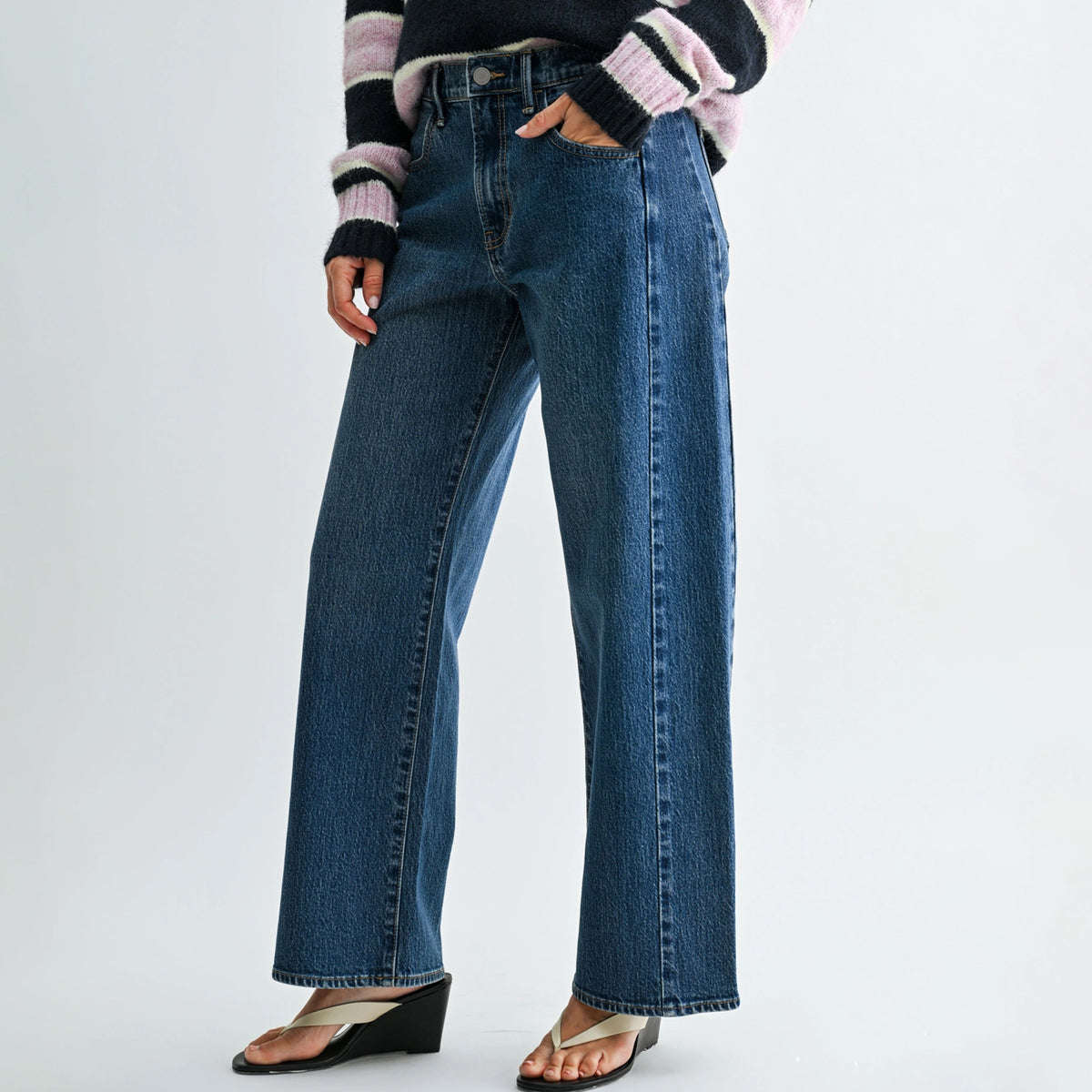 A person wearing high-waisted, wide-leg blue denim jeans with a striped knit sweater and wedge sandals, shown from the chest down against a plain light gray studio background.