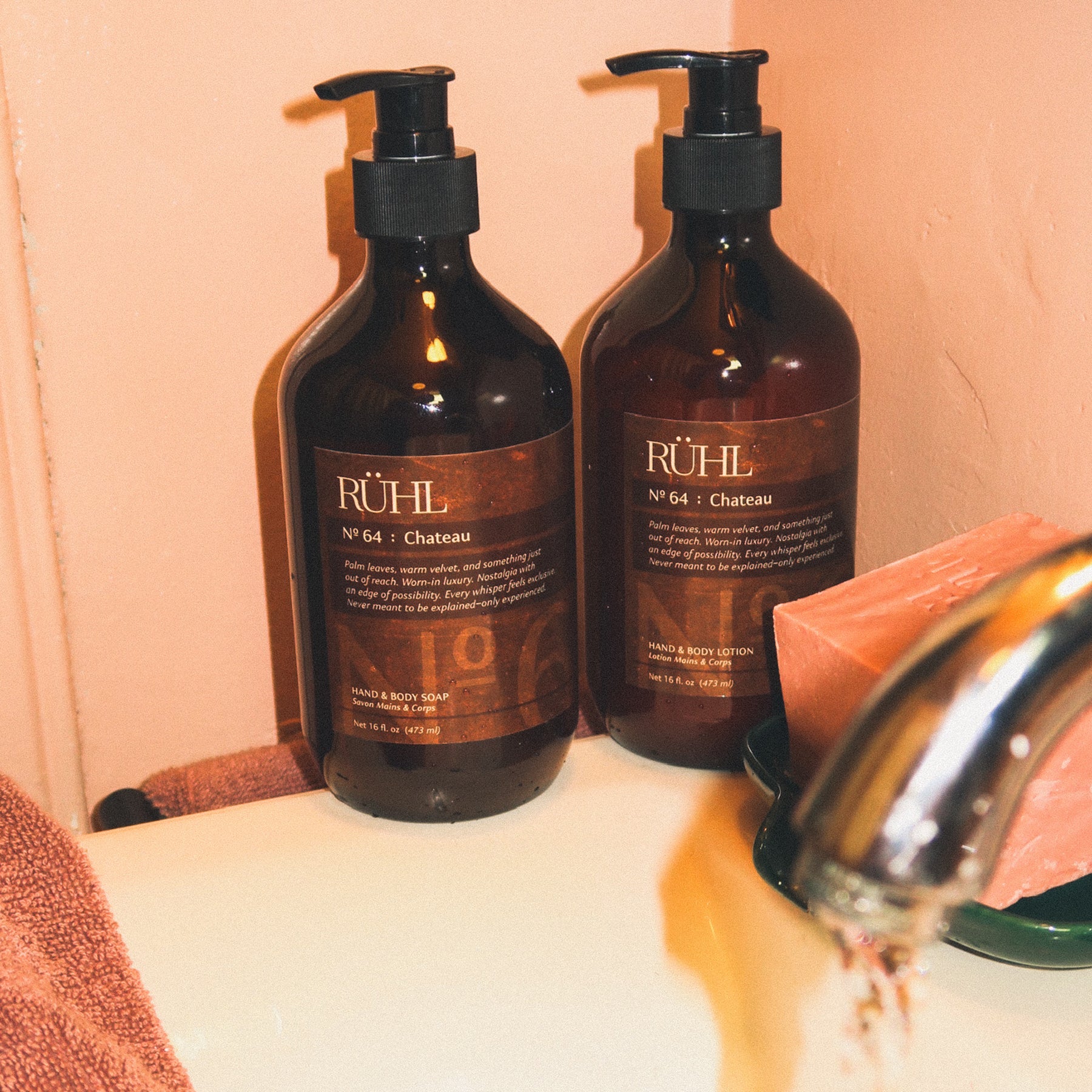 Two amber pump bottles of Rühl No. 64 Chateau body wash and hand wash standing on a bathroom counter next to a running faucet, with a pink soap bar and towel visible.