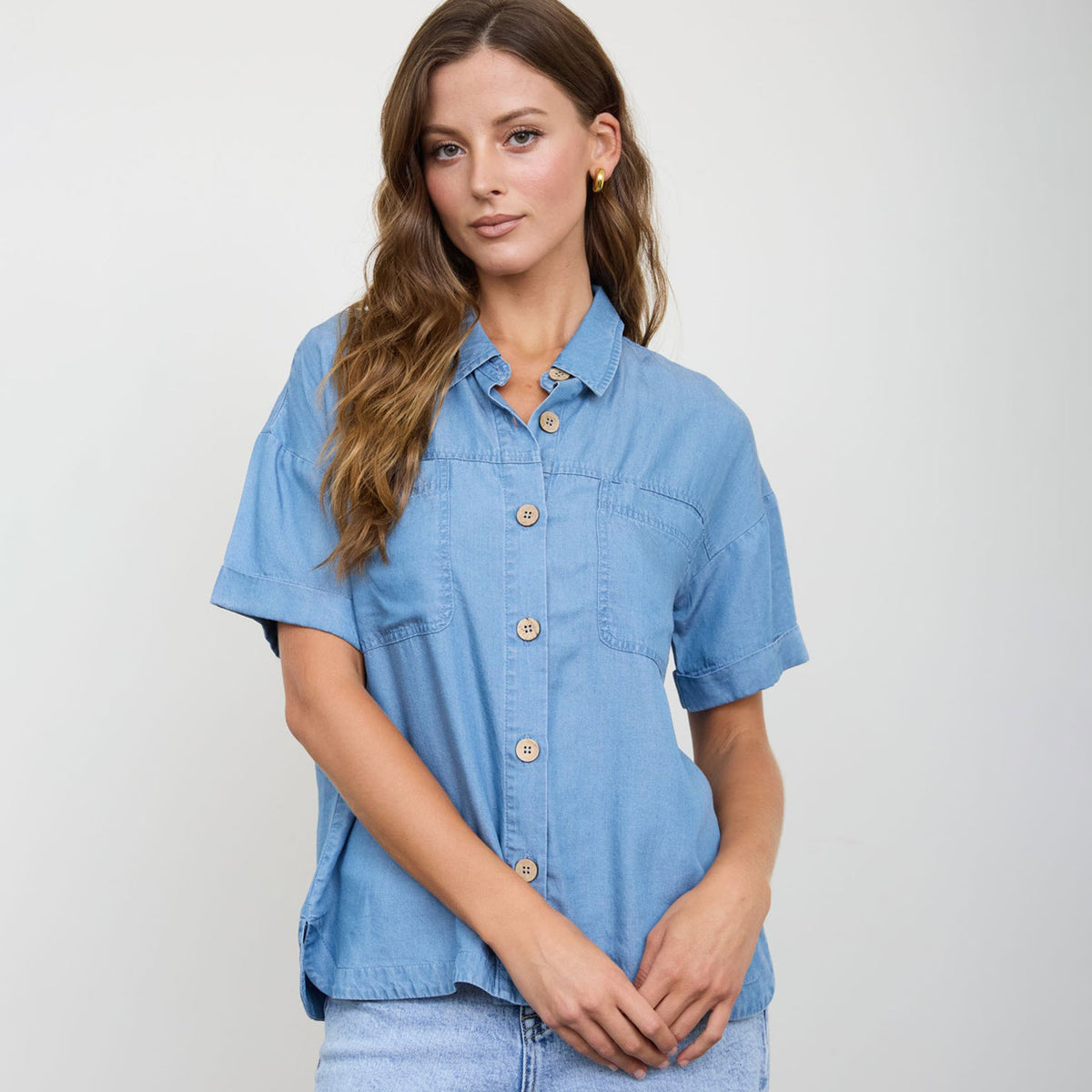 Woman modeling a button down chambray top with short sleeves and front pocket detail.