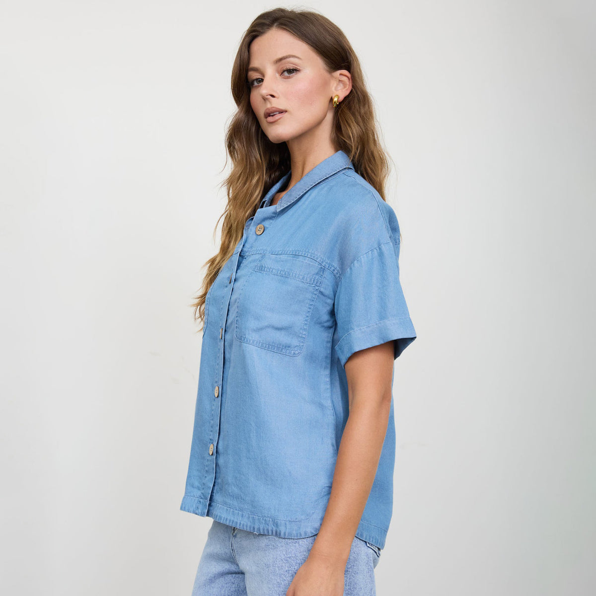 Woman modeling the side view of a button down chambray top with short sleeves and front pocket detail.
